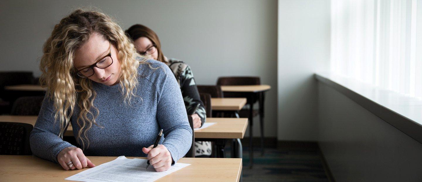 Two female students seated at desks, writing on documents.